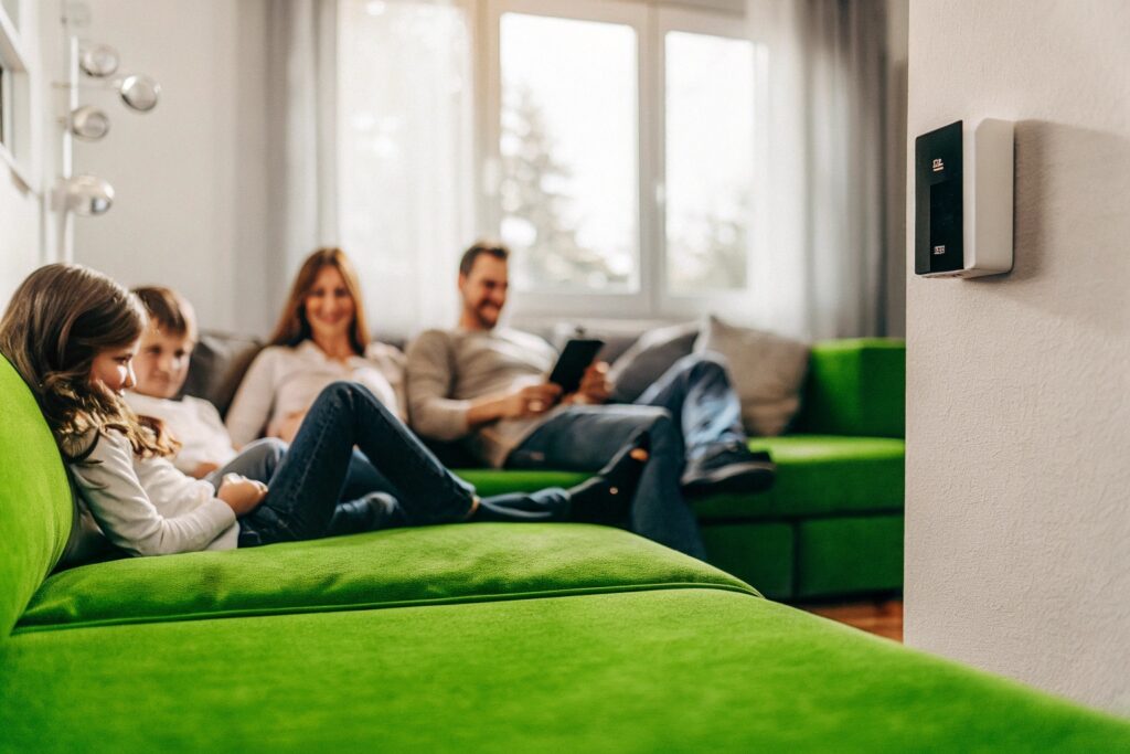 Familie entspannt im Wohnzimmer mit moderner Smart-Home-Steuerung an der Wand – intelligente Gebäudetechnik sorgt für Komfort, Sicherheit und Energieeffizienz im Alltag.