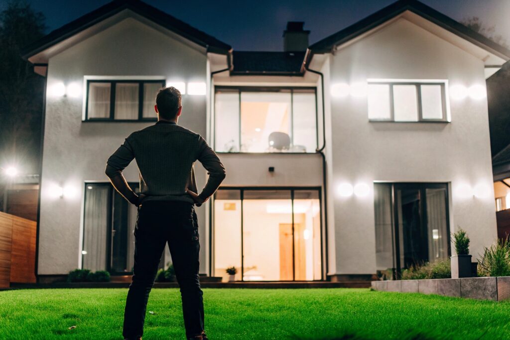 Hausbesitzer steht vor einem modern beleuchteten Smart Home bei Nacht – smarte Außenbeleuchtung sorgt für Sicherheit, Komfort und Energieeffizienz.