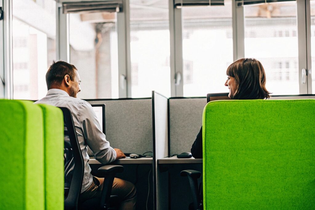 Zwei Mitarbeitende arbeiten in einem modernen Büro mit abgetrennten Arbeitsplätzen und großen Fenstern. Die Szene steht beispielhaft für eine gut klimatisierte Arbeitsumgebung mit Fokus auf Komfort und Konzentration.