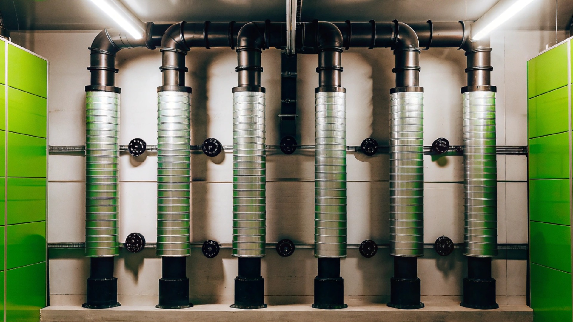 Sechs vertikale Lüftungsrohre mit schwarzem Leitungssystem in einem Technikraum, flankiert von grünen Wandverkleidungen. Sie sind Teil eines modernen Versorgungstechniksystems.